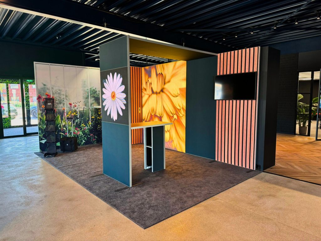 Een kleurrijke tentoonstellingsruimte met grote bloemmotieven, inclusief een paneel met levendige bloemen en een display met echte planten, in een moderne ruimte met houten vloeren en een donker plafond.