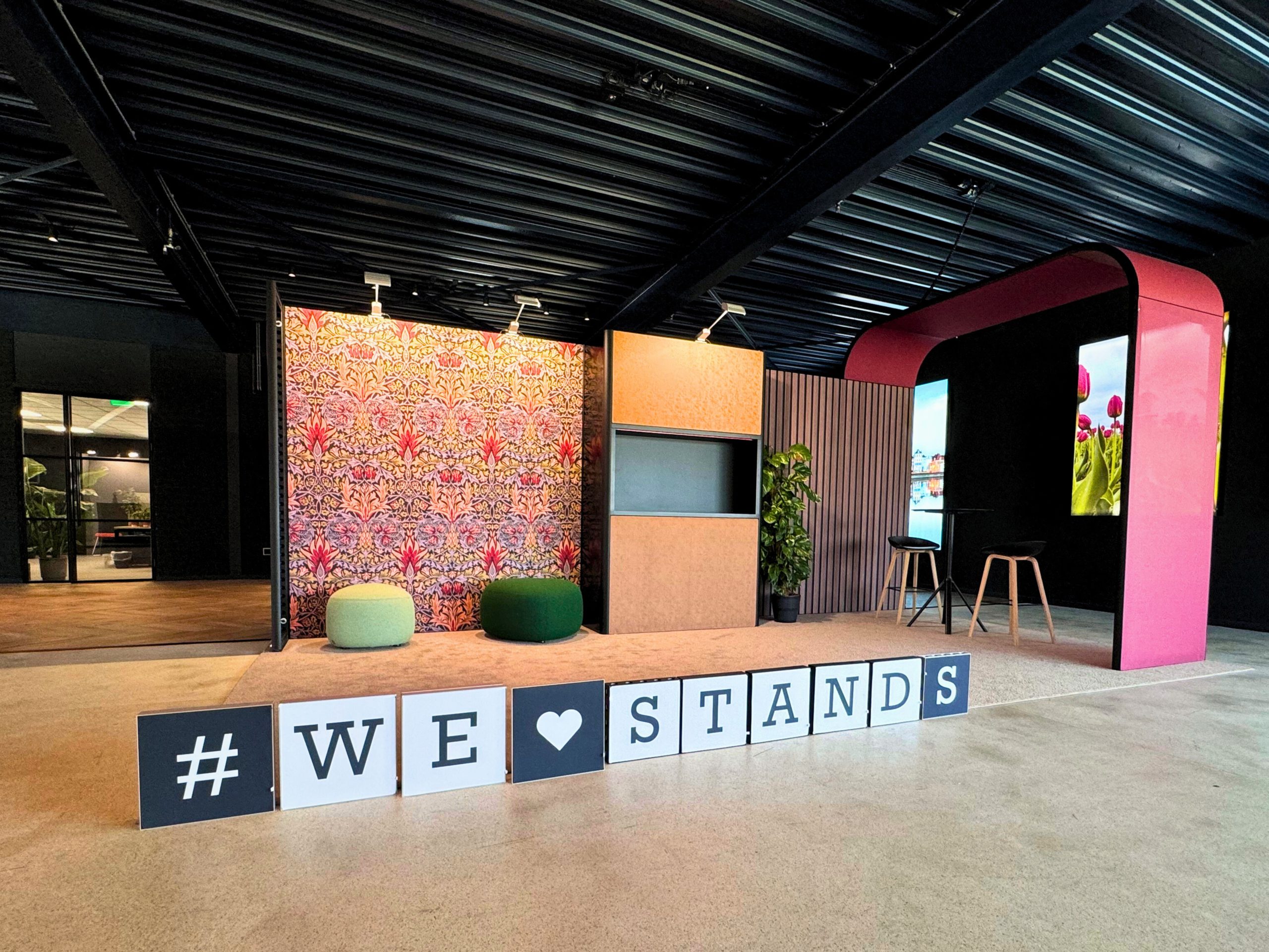 Een stijlvol ingerichte beursstand met een kleurrijke achtergrond, moderne meubels en een hashtag "#WE ❤️ STANDS" in een ruime hal met een donker plafond.