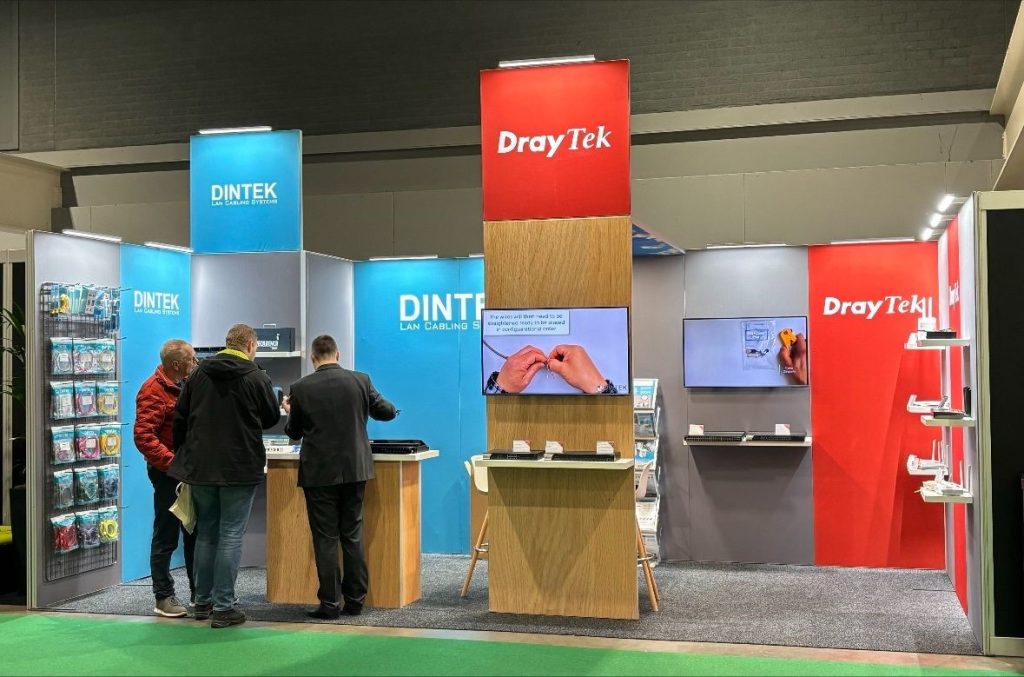 Een beursstand met 3 mensen bij DINTEK en DrayTek, waar bezoekers in gesprek zijn met vertegenwoordigers, omringd door technologieproducten en displays.